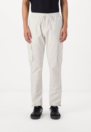 Pantalons cargo beiges clairs avec une taille élastique, fermeture à cordon de serrage, deux poches latérales et une texture lisse, assortis à des baskets noires.