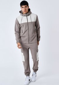 Lichtbruine zip-up hoodie met witte panelen, voorzien van trekkoorden. Bijpassende joggers hebben grote zijzakken en ribgebreide boorden. Witte sneakers.