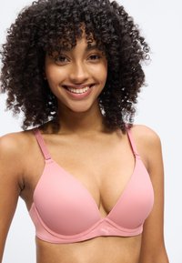 Χαμογελαστή γυναίκα με σγουρά μαλλιά που φορά ροζ επενδεδυμένο σουτιέν με λεπτές ρυθμιζόμενες τιράντες, μπροστά από απλό λευκό φόντο.