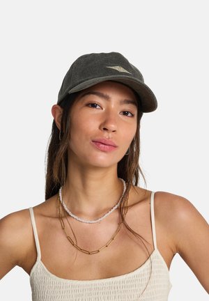 Casquette en coton vert foncé à visière incurvée avec un petit logo à l'avant, portée par une personne aux cheveux longs et portant plusieurs colliers superposés.