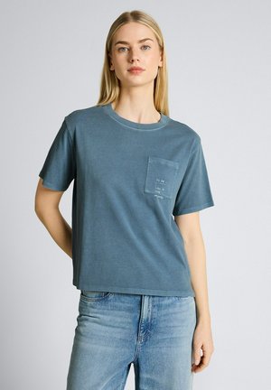 Młoda kobieta o długich blond włosach w niebieskiej koszulce z przednią kieszenią i jasnoniebieskich dżinsach stoi na jednolitym tle.