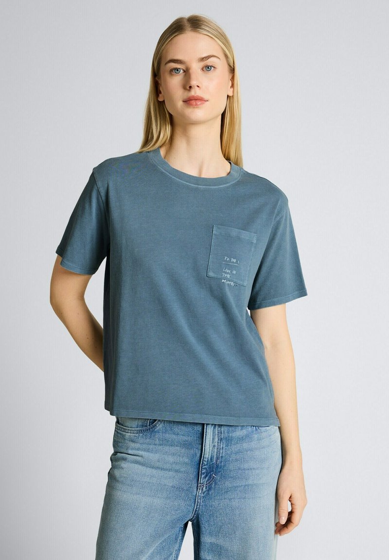 Junge Frau mit langen blonden Haaren trägt ein blaues T-Shirt mit Brusttasche und hellblaue Jeans, steht vor einem einfarbigen Hintergrund.