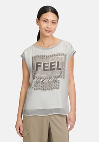 Haut gris clair à manches courtes avec un motif texturé sur le devant présentant le mot "FEEL" en lettres 3D, des accents floraux et des détails cloutés. Tissu doux.
