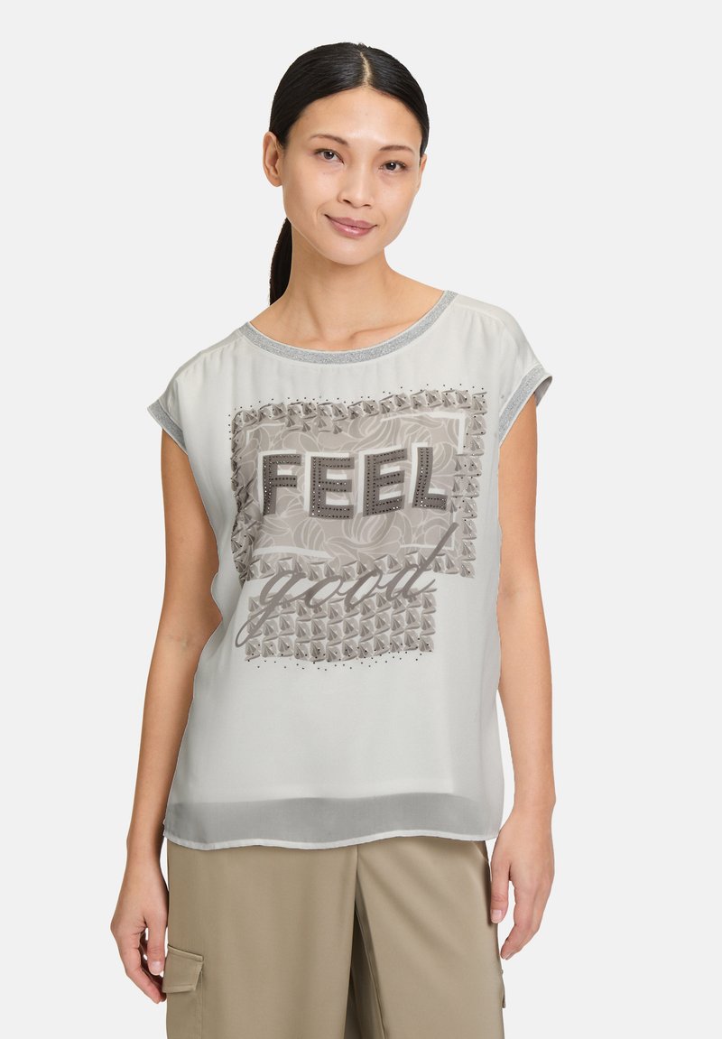 Haut gris clair à manches courtes avec un motif texturé sur le devant présentant le mot "FEEL" en lettres 3D, des accents floraux et des détails cloutés. Tissu doux.