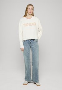 Maglione bianco a coste con la scritta "TRUE RELIGION" in colore pesca, abbinato a jeans larghi celeste chiaro e sneakers bianche con suola beige.