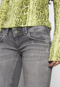 Grå denimjeans har en smal passform, distinkta fickkonturer och gröna knappar. En ljus gulgrön ormskinnstrykt topp har texturerad detaljering.