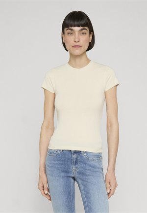 Femme aux cheveux courts et foncés portant un haut crème ajusté à manches courtes et un jean en denim bleu clair, debout devant un fond gris uni.