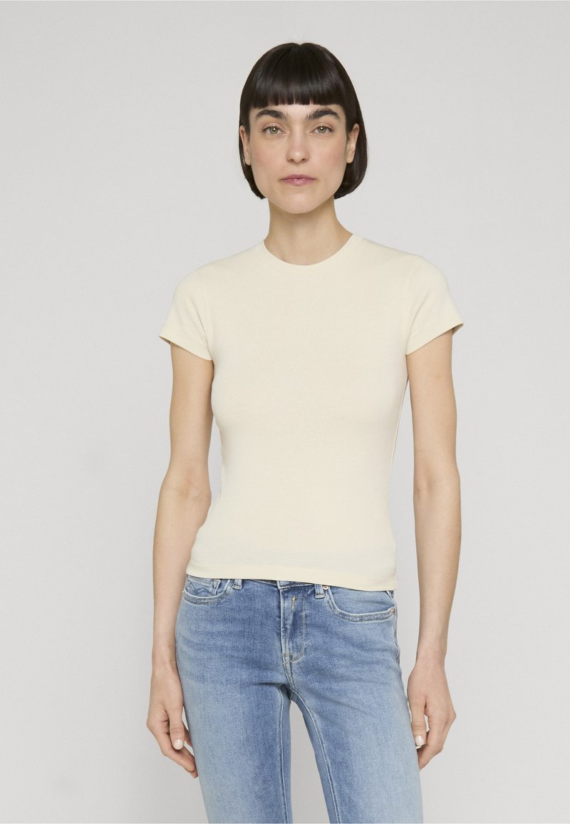 Femme aux cheveux courts et foncés portant un haut crème ajusté à manches courtes et un jean en denim bleu clair, debout devant un fond gris uni.