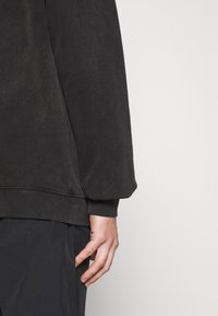 Felpa grigia scura, caratterizzata da un collo rotondo e maniche lunghe con polsini a costine, esposta insieme a pantaloni neri.