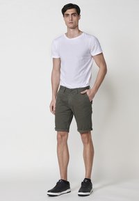 Tricou alb cu guler rotund, pantaloni scurți khaki cu mansete și adidași negri. Ținuta are un design casual, ajustat, cu detalii minime.