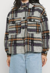 Geruite jas van zachte stof, met grijze, zwarte, paarse en oranje kleuren. Voorzien van twee borstzakken en zwarte knopen.