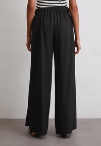 Pantalons larges noirs en tissu texturé, dotés d'une taille élastique et d fentes latérales à l'ourlet, vus de dos.