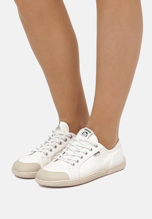 Personne portant des baskets basses blanches avec des bouts et des semelles beiges, des lacets blancs et le logo « mtng » sur la languette et le côté.