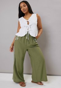 Vit ärmlös blus med scalloped kanter och snörning framtill, kombinerad med olivgröna vida byxor med blommönster broderat på fickorna.