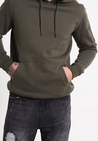Felpa verde oliva realizzata in tessuto morbido, con tasca a marsupio, coulisse regolabile e tasca con zip sulla manica. Indossata con jeans scuri.