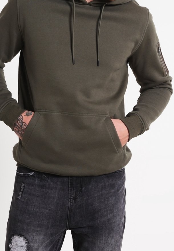 Felpa verde oliva realizzata in tessuto morbido, con tasca a marsupio, coulisse regolabile e tasca con zip sulla manica. Indossata con jeans scuri.
