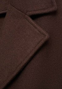Dettaglio di un cappotto di lana marrone con una finitura morbida e testurizzata. Presenta un colletto a ribelle e rever strutturati.
