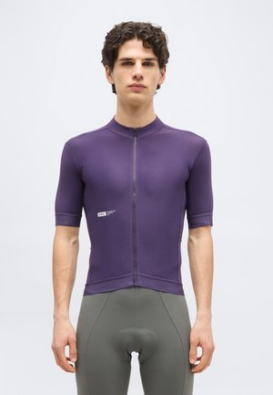 Νέος άνδρας όρθιος, φορώντας εφαρμοστή μοβ φανέλα ποδηλασίας με κοντά μανίκια και εμπρόσθιο φερμουάρ, καθώς και γκρι επενδυμένα σορτς ποδηλασίας.