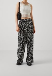 Žena v bílém krajkovém topu bez rukávů a širokých černobílých kalhotách, černých teniskách a černé tašce přes rameno.