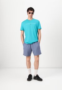 Turquoise katoenen t-shirt met "TOMMY JEANS" logo, grijze korte broek, zwarte sneakers, witte sokken, ronde zonnebril, staand tegen een effen achtergrond.