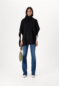 Poncho en tricot noir avec un col haut en côte, associé à un jean bleu évasé. Le modèle tient un sac vert, se tenant contre un fond uni.