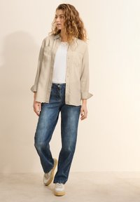 Camisa beige de botones sobre una camiseta blanca, combinada con jeans a rayas azules y zapatillas blancas. La camisa tiene dos bolsillos en el pecho.