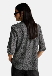 Czarna koszula z małym białym wzorem kwiatowym, długie rękawy podwinięte, kołnierz i luźny krój, pokazana z tyłu.