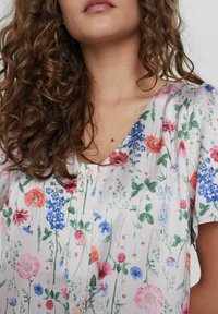 Top con motivo floreale in tessuto leggero; presenta fiori rosa, blu e rossi su sfondo bianco; scollatura a V con bottoni piccoli.
