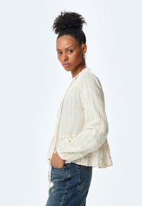 Blusa beige chiaro con righe verticali bianche, orlo a peplo, abbottonatura frontale, maniche lunghe e tessuto morbido e testurizzato; abbinata a jeans blu.