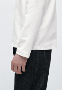 Weißes Langarmshirt mit Rundhalsausschnitt und glatter Textur. Kombiniert mit dunklen Jeanshosen, die ein schlankes Design zeigen.