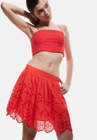 Top corto rosso con ricami intricati e spalline regolabili, abbinato a una gonna lunga fino al ginocchio con dettagli floreali ritagliati e una vita elasticizzata.