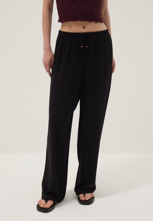 Donna che indossa pantaloni neri larghi con coulisse, top corto lavorato a maglia color bordeaux e sandali platform neri, in piedi su uno sfondo chiaro e uniforme.