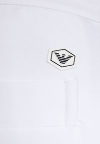 Tela blanca con una textura suave, que presenta un parche de logo hexagonal en negro con líneas estilizadas, ubicado en el área del bolsillo.