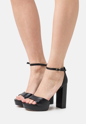 Sandales à talons hauts en cuir noir avec une large bride sur les orteils et une bride à la cheville ornée d'une boucle dorée sur un talon bloc.