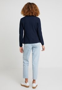 Granatowa marynarka w groszki o dopasowanym kroju, zestawiona z jasnoniebieskimi jeansami. Białe trampki mają fakturowaną podeszwę, dopełniając stylizację.