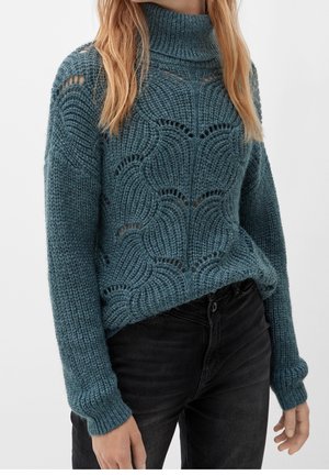 Femme portant un pull à col roulé bleu en maille avec un motif ondulé et un jean noir, debout devant un fond uni.