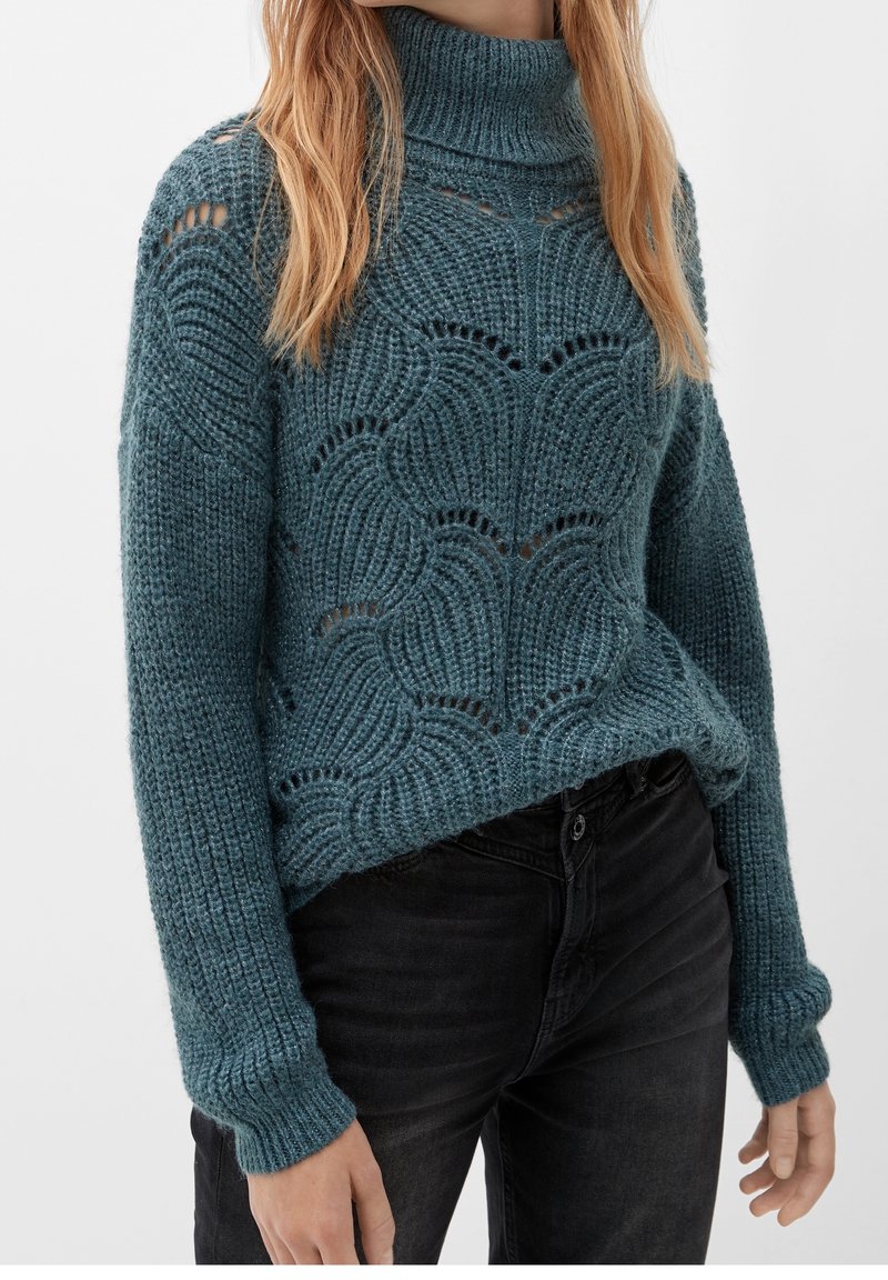 Femme portant un pull à col roulé bleu en maille avec un motif ondulé et un jean noir, debout devant un fond uni.