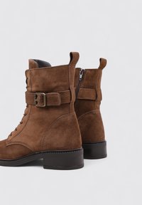 Botines de ante marrón hasta el tobillo con punta redondeada, parte delantera con cordones, una correa con hebilla y cremallera lateral. Tacón grueso para mayor soporte.