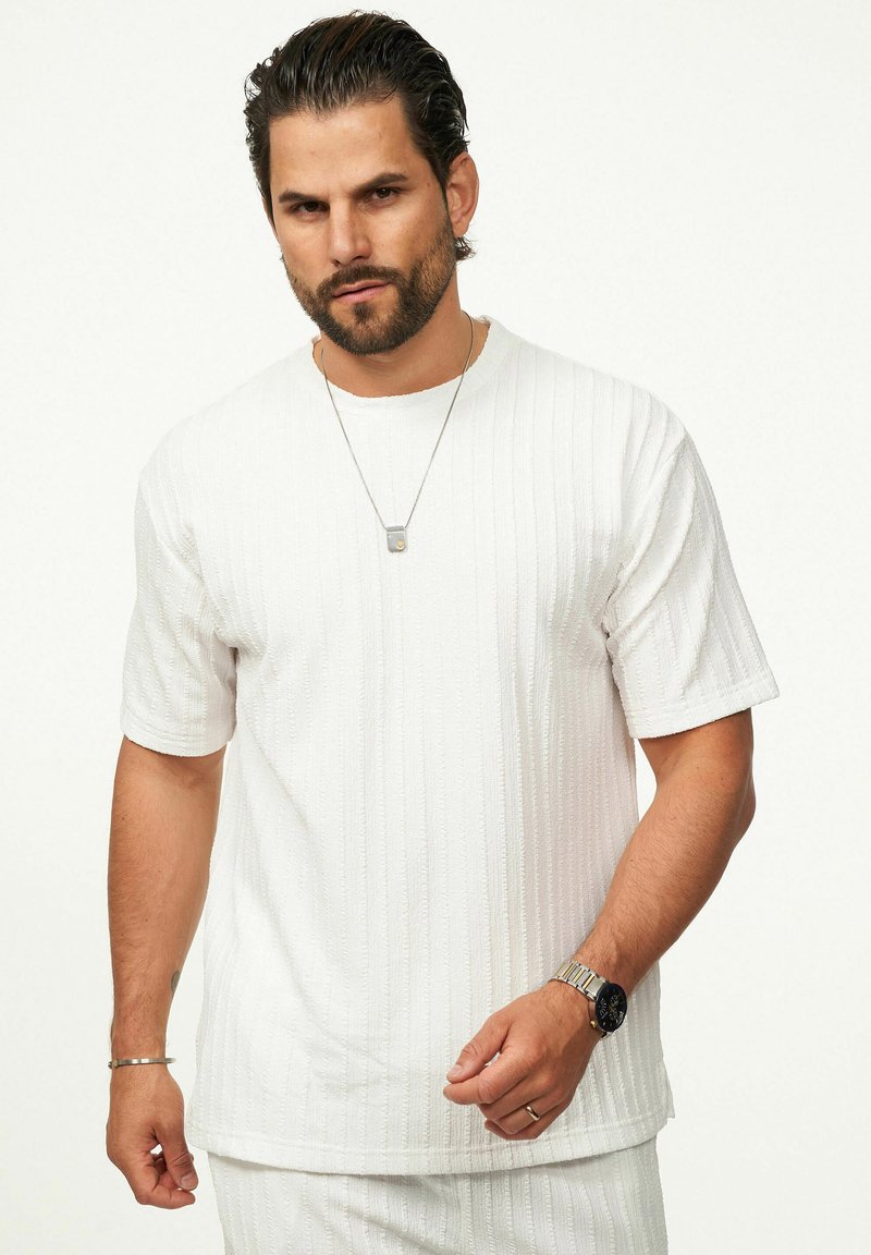 White short-sleeve shirt with textured vertical stripes, round neckline, and relaxed fit. Silver necklace and wristwatch visible.
