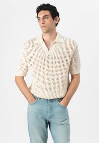 Suéter de polo de manga corta de punto en color crema con un patrón de tejido abierto. Combinado con unos jeans ajustados de color azul claro, con las manos en los bolsillos.