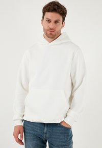 Pehmeästä kankaasta valmistettu valkoinen huppari, jossa on etutasku ja resorit hihansuissa, yhdistetty sinisiin farkkuihin.