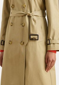 Beige trench coat dengan kancing ganda, ikat pinggang di pinggang, dan tekstur yang menarik. Mencakup aksen merah yang kontras di lengan.