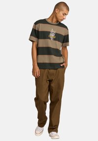 Camiseta a rayas en verde oscuro y beige, con un gráfico de llama y rosa. Combinada con pantalones marrones y zapatillas blancas.