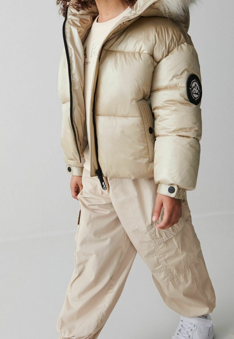 Enfant portant une doudoune beige avec capuche en fourrure, pantalon cargo beige et baskets blanches, debout sur un fond uni.