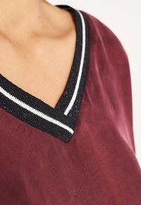 Burgundy top with a V-neck, featuring a black and white striped ribbed collar with a subtle shimmer and smooth texture.