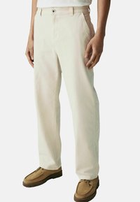 Light beige cotton pants with a straight-leg cut, featuring side pockets and a button closure, paired with brown suede shoes.