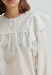 Kobieta w białej bluzce z małym zapięciem na guziki, marszczonym materiałem poniżej biustu i dużymi falbaniastymi rękawami.