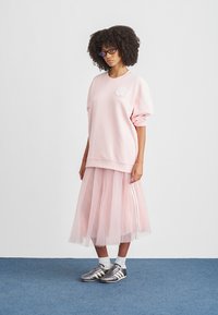 Růžová mikina s texturovaným logem, kombinovaná s průsvitnou růžovou tylovou sukní s bílými pruhy, doplněná šedými sportovními botami.