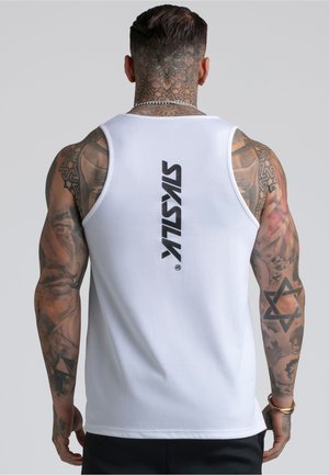 Gespiierde, getatoeëerde man die een wit mouwloos shirt draagt met het "SikSilk"-logo verticaal gedrukt op de achterkant, staand tegen een effen achtergrond.