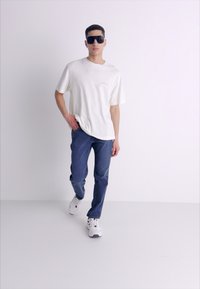 Camiseta blanca de cuello redondo combinada con jeans azules; el modelo lleva gafas de sol negras y zapatillas blancas; un conjunto elegante y casual.